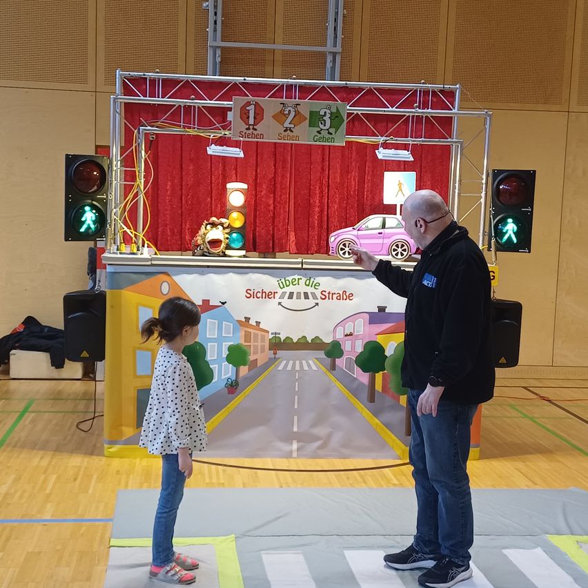 An adult man and a young girl stand in front of a stage with a cartoon street scene. The adult points to the scene while the girl looks at it.