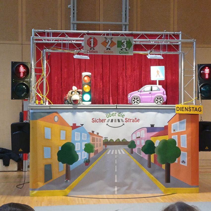 A stage setup with a red curtain and a small car on it, traffic lights, and a stage backdrop with buildings and trees. The backdrop reads 'Sicher uber die Strasse'.