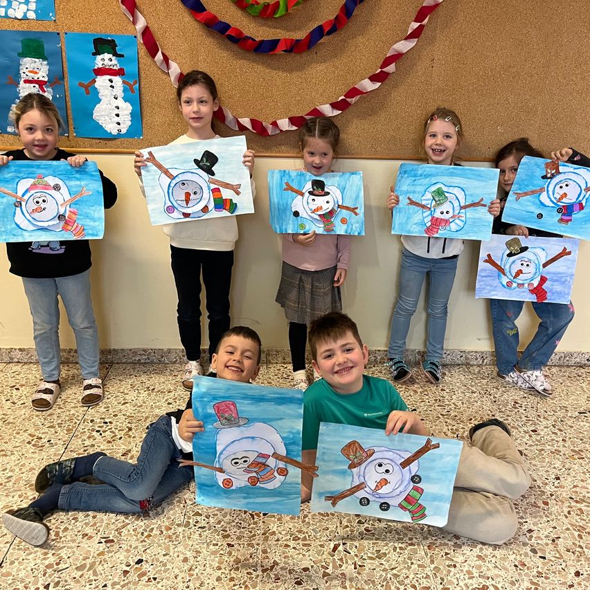 Eine Gruppe von Kindern ist drinnen, lächelt und hält Gemälde von Schneemännern. Sie sind warm gekleidet und einige tragen Schuhe. Die Wand hinter ihnen ist mit bunten Bändern und Schneemanngemälden dekoriert.