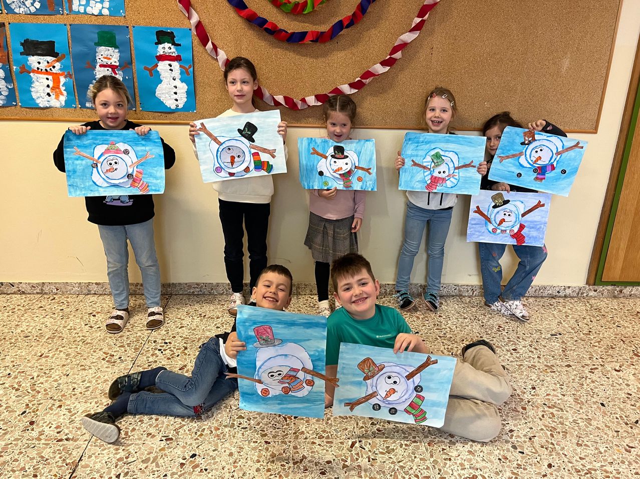Eine Gruppe von Kindern ist drinnen, lächelt und hält Gemälde von Schneemännern. Sie sind warm gekleidet und einige tragen Schuhe. Die Wand hinter ihnen ist mit bunten Bändern und Schneemanngemälden dekoriert.