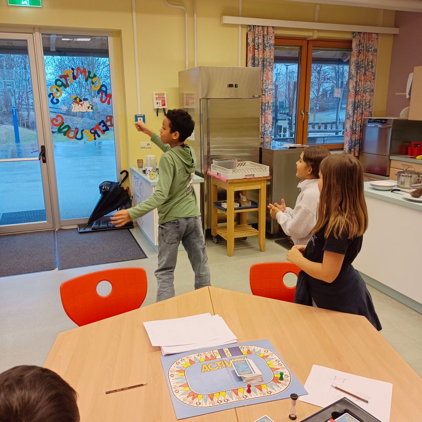 Drei Kinder befinden sich in einem Raum mit einem Brettspiel auf dem Tisch. Ein Junge zeigt auf eine Glastür mit bunten Dekorationen.