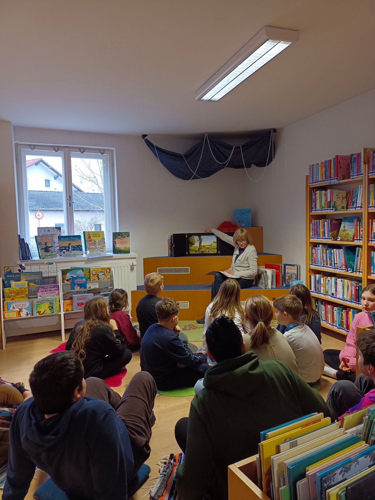 Ein Klassenzimmer mit Schülern, die in einem Kreis um eine Frau sitzen, die auf einer Bühne liest, umgeben von Bücherregalen und einem Fenster mit Blick auf ein Haus.