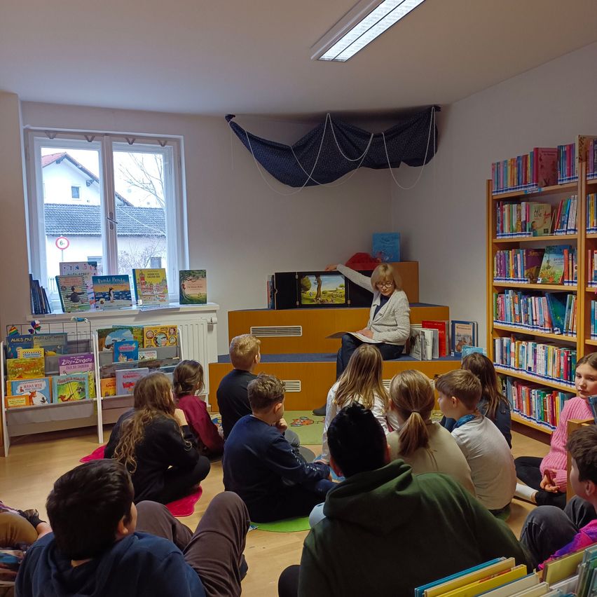 Ein Klassenzimmer mit Schülern, die in einem Kreis um eine Frau sitzen, die auf einer Bühne liest, umgeben von Bücherregalen und einem Fenster mit Blick auf ein Haus.