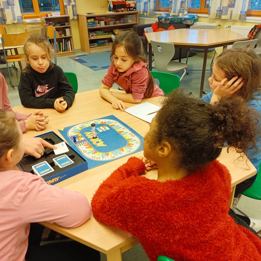 Fünf Mädchen sitzen um einen Tisch und spielen ein Brettspiel in einem Klassenzimmer. Ein blaues Brett mit verschiedenen farbigen Kreisen liegt auf dem Tisch. Sie sind tief in das Spiel vertieft.