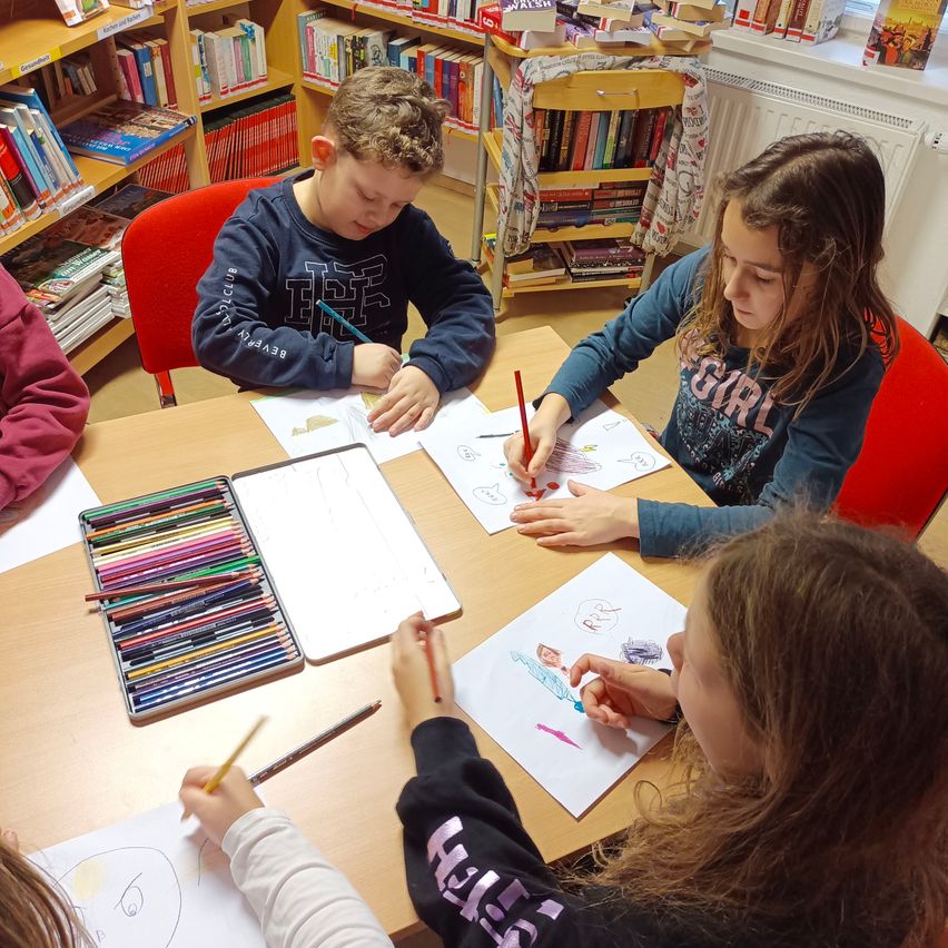 In einer Bibliothek sitzen eine Gruppe von Kindern um einen Tisch. Sie färben eine Zeichnung mit Buntstiften ein.
