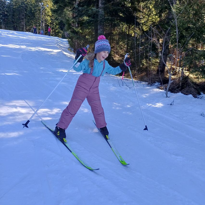 Ein junges Mädchen schreitet einen verschneiten Hang hinunter, in einer blauen Jacke, pinken Hosen und einer Mütze. Sie hält Skistöcke und lächelt. Bäume und eine entfernte Figur sind im Hintergrund zu sehen.