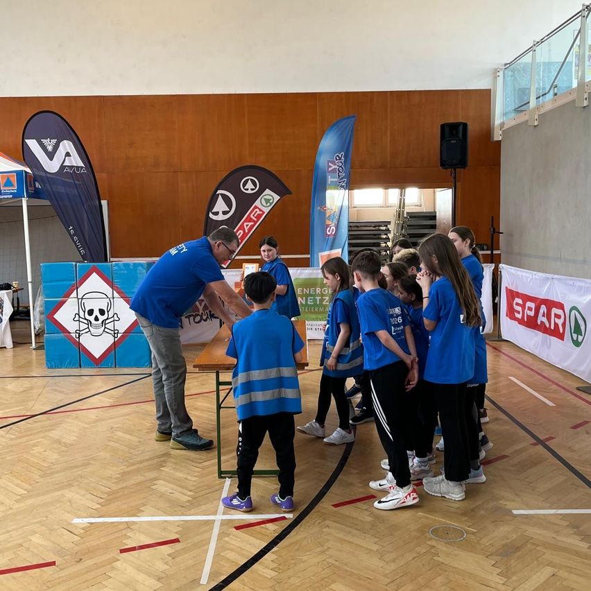 Ein Mann in einem blauen Shirt überreicht einem Jungen in einer blauen Weste eine Sicherheitsauszeichnung. Mehrere Kinder stehen hinter ihnen auf einem Basketballplatz.