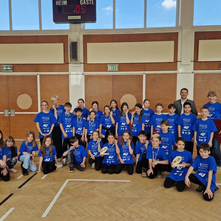 Eine Gruppe junger Personen in blauen Shirts und schwarzen Hosen posiert für ein Foto in einem Innenbereich. Sie tragen alle Medaillen um den Hals. Das an der Wand montierte Spielfeld zeigt den Spielstand an.