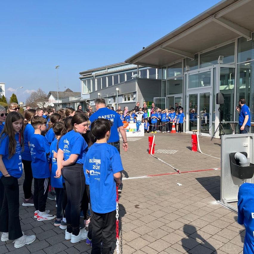 Eine Gruppe von Kindern in blauen T-Shirts mit verschiedenen Nummern und Texten steht in einer Schlange vor einem Gebäude mit Glaswänden und einer Person in der Nähe eines Lautsprechers.