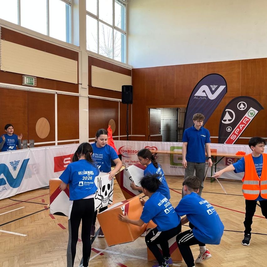 Mehrere Schüler in blauen Uniformen sind in einer Turnhalle mit einer Aktivität beschäftigt. Sie arbeiten an einer orangefarbenen Struktur, und einige haben Sicherheitswesten. Banner mit Logos sind im Hintergrund.