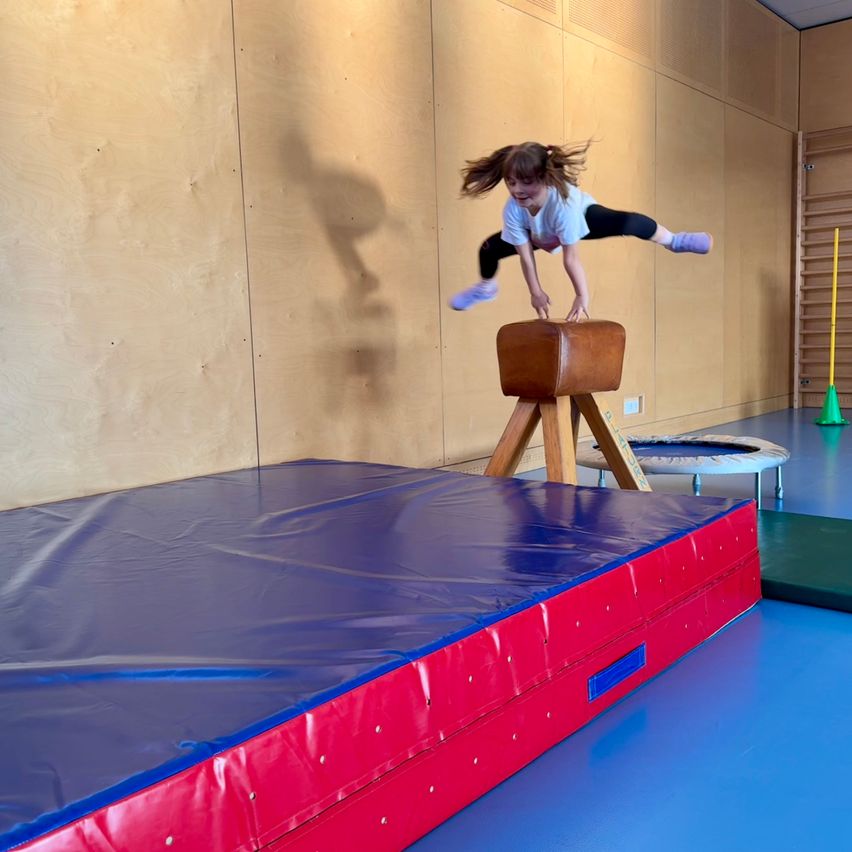 Ein junges Mädchen führt einen Handstand auf einem hölzernen Sprungpferd in einem Fitnessstudio aus. Der Boden der Turnhalle ist blau und vor ihr ist eine blaue Matte ausgelegt.