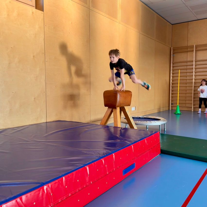 Ein junger Junge springt über ein gepolstertes Hindernis in einer Turnhalle. Ein weiteres Kind steht in der Nähe, und ein Trampolin und ein gelber Pfahl sind im Hintergrund zu sehen.