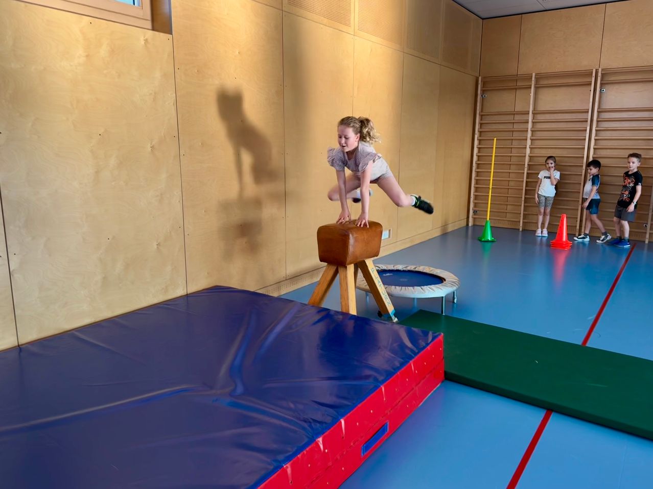 Ein kleines Mädchen springt in einem Fitnessstudio über einen Barren, mit einem Trampolin und Kegel im Hintergrund.
