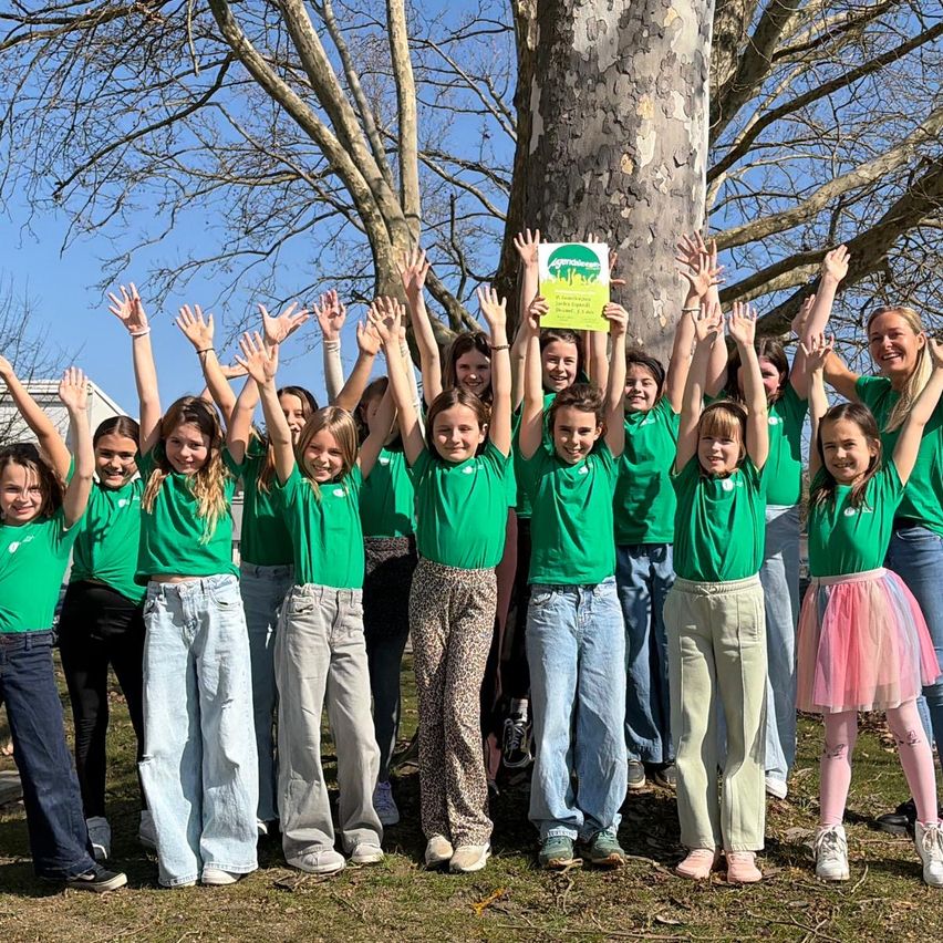 Eine Gruppe junger Mädchen und eine Frau in grünen Shirts stehen unter einem Baum und halten ein gelbes Schild. Sie lächeln und heben ihre Hände.