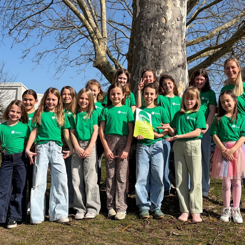 Eine Gruppe von Kindern in grünen T-Shirts posiert für ein Foto mit einem Baum. Sie sind draußen an einem sonnigen Tag.