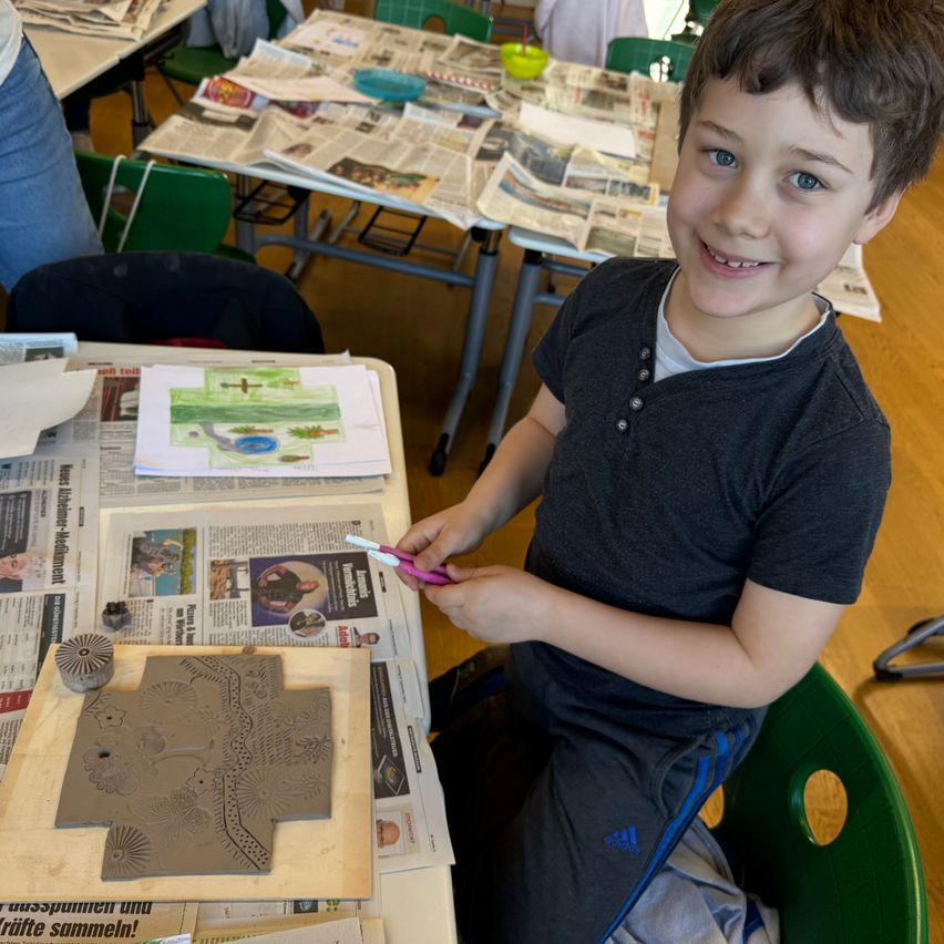 Ein junger Junge in einem Klassenzimmer benutzt eine Schere, um Ton zu schneiden. Er sitzt an einem mit Zeitungspapier und Ton bedeckten Tisch.