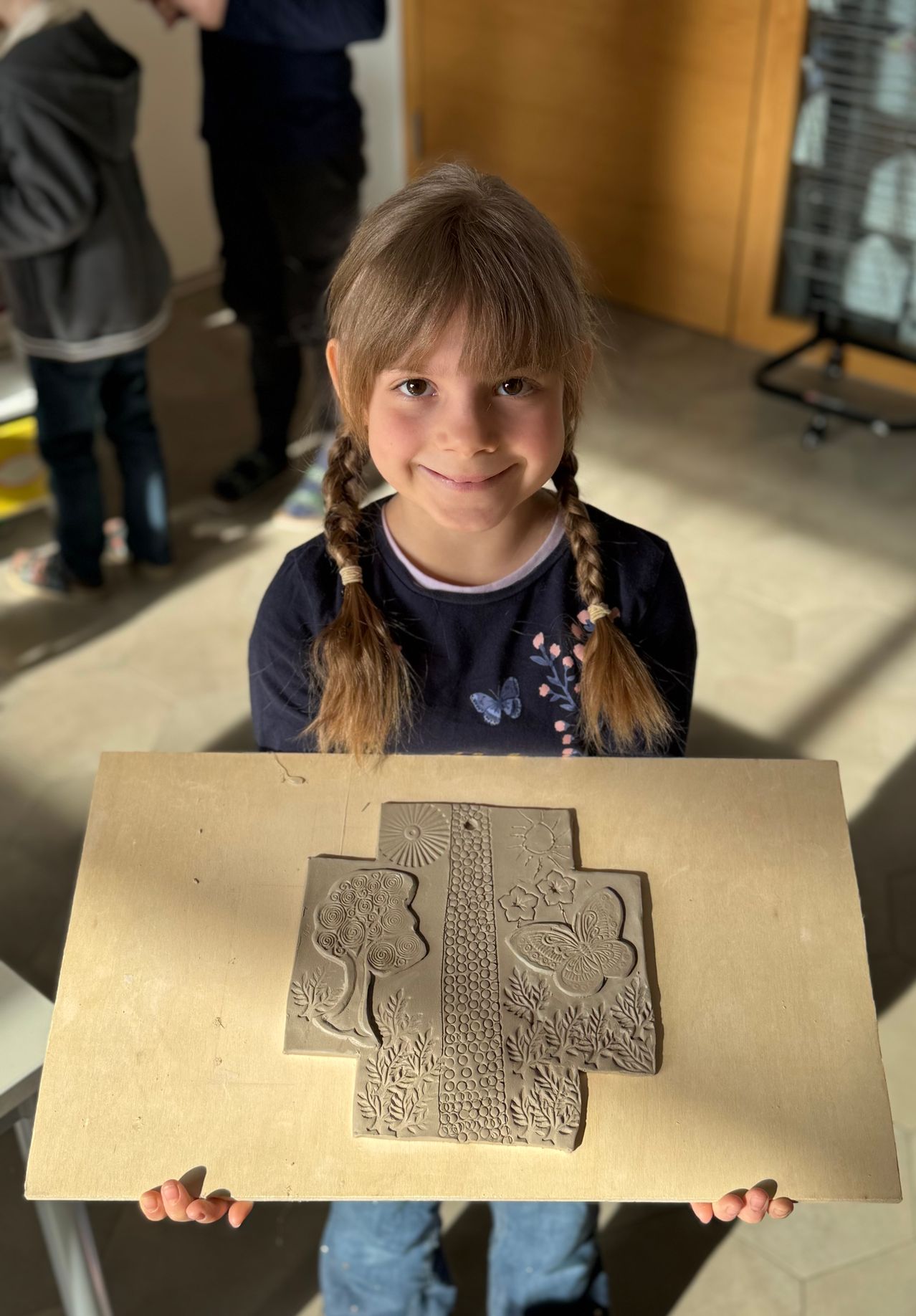 Ein kleines Mädchen mit geflochtenen Haaren lächelt und posiert für ein Foto mit einem Tonkunstwerk auf einem Tisch vor ihr.