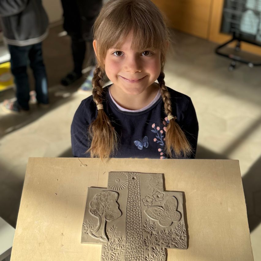 Ein kleines Mädchen mit geflochtenen Haaren lächelt und posiert für ein Foto mit einem Tonkunstwerk auf einem Tisch vor ihr.