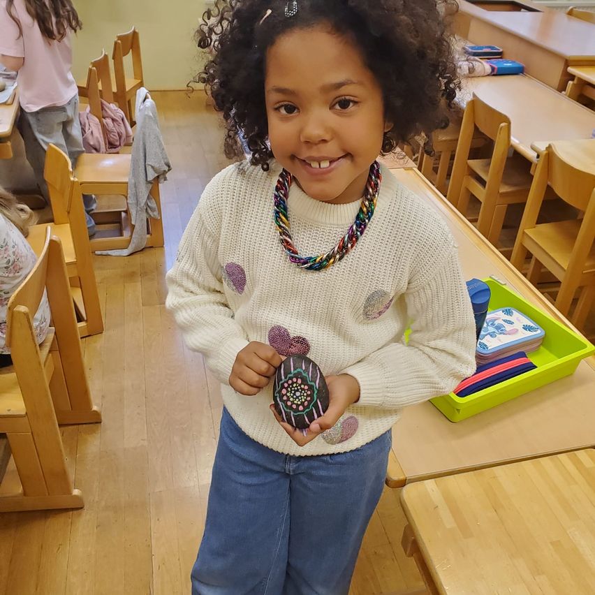 Ein lächelndes kleines Mädchen mit lockigem Haar steht im Klassenzimmer und hält einen dekorativen Stein. Sie trägt einen weißen Pullover und blaue Jeans. Hinter ihr sind Tische angeordnet, und ein gelbes Tablett hält verschiedene Gegenstände.