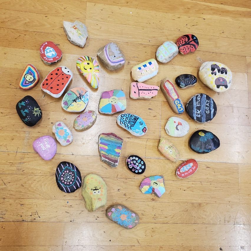 Eine Sammlung bemalter Steine, die auf einem Holzboden angeordnet sind und verschiedene Designs wie eine Marienkäferin, eine Wassermelone und eine Sonne zeigen.