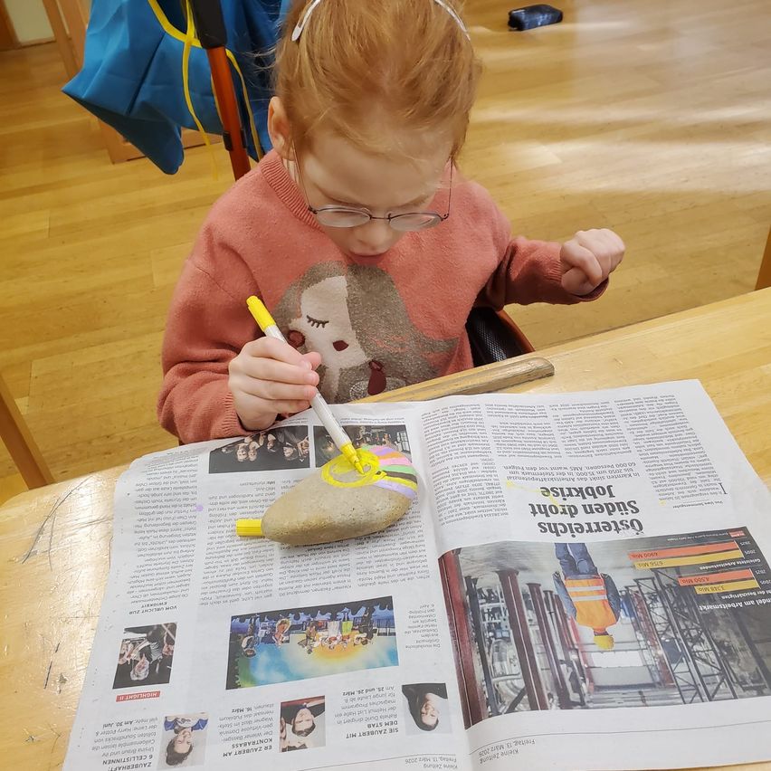 Ein junges Kind, das eine Brille und einen rosa Pullover trägt, malt einen Stein mit einem Marker auf einer Zeitung. Die Zeitung hat ein Bild eines Mannes in einer Sicherheitsweste.