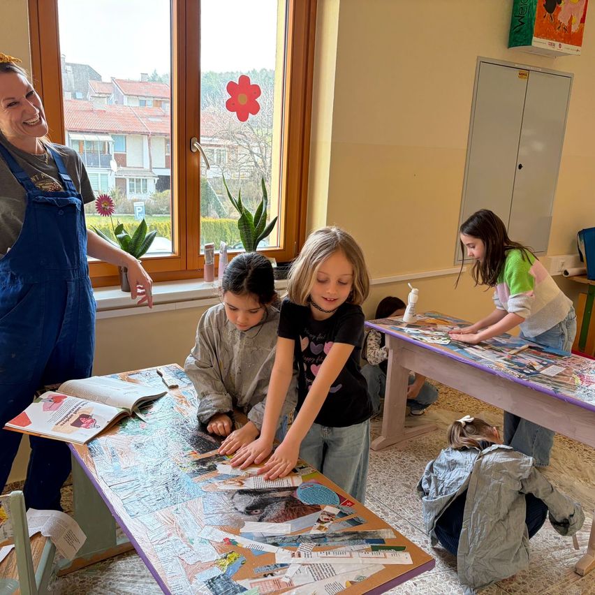 Eine Gruppe von Kindern und einem Erwachsenen arbeitet in einem Klassenzimmer an einem Projekt. Die Kinder sitzen an einem Tisch und arbeiten an einer Collage mit verschiedenen Papierstücken. Die Frau steht und lächelt. Es gibt Pflanzen und Blumen auf der Fensterbank.