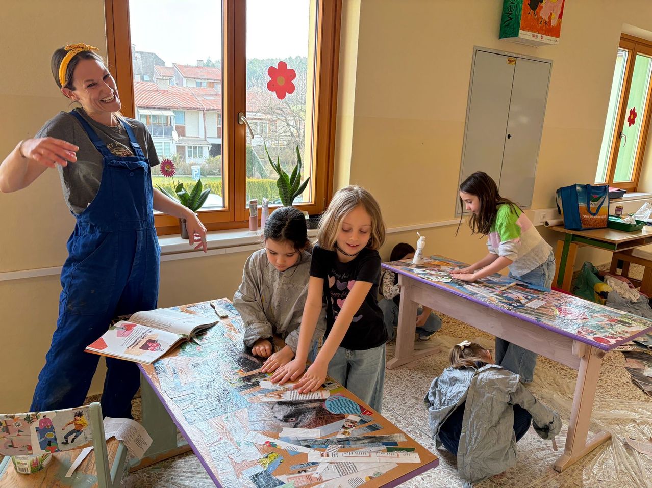 Eine Gruppe von Kindern und einem Erwachsenen arbeitet in einem Klassenzimmer an einem Projekt. Die Kinder sitzen an einem Tisch und arbeiten an einer Collage mit verschiedenen Papierstücken. Die Frau steht und lächelt. Es gibt Pflanzen und Blumen auf der Fensterbank.
