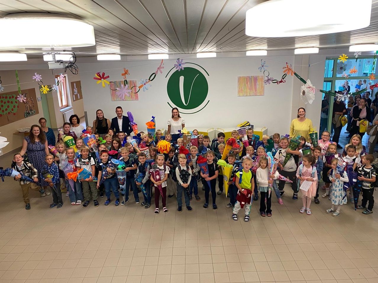 Eine Gruppe von Kindern und Erwachsenen versammelt sich in einem Raum, hält Geschenke und lächelt für ein Foto. Der Raum ist mit bunten Blumen und einem grünen Logo an der Wand dekoriert.