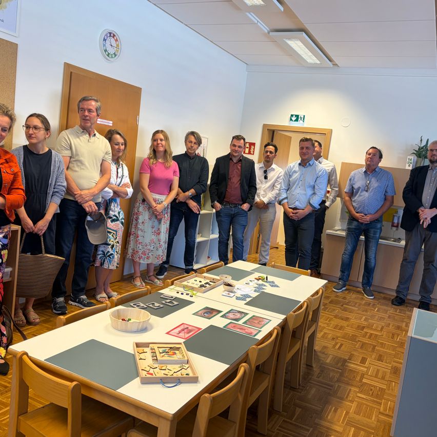 Eine Gruppe von Menschen steht um einen Tisch in einem Raum. Sie schauen sich Puzzles auf dem Tisch an. Der Raum hat einen Holzboden und eine Uhr an der Wand.