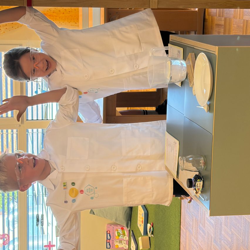 Zwei Kinder in weißen Laborkittel posieren für ein Foto. Sie tragen Brillen und scheinen ein wissenschaftliches Experiment durchzuführen. Ein Tisch mit einem Teller, einem Krug und einer Tintenflasche ist im Hintergrund zu sehen.