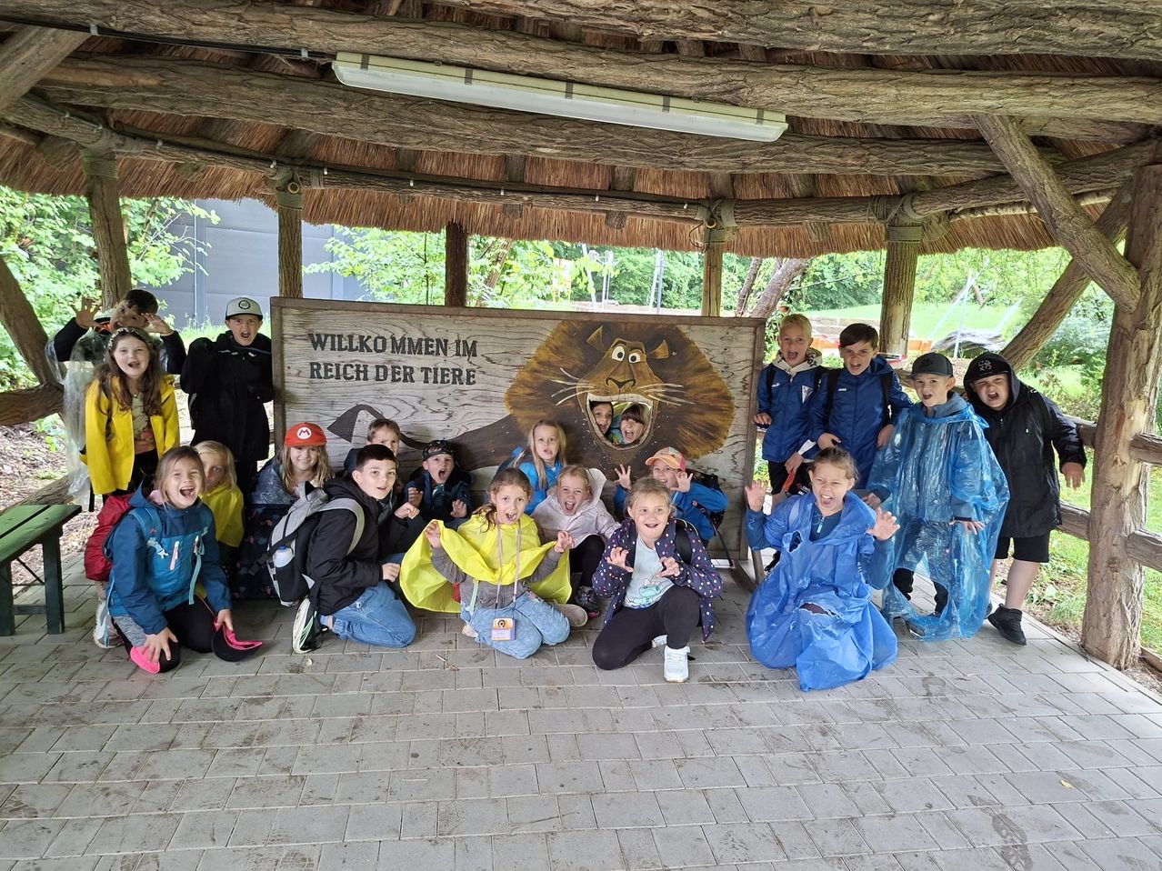 A group of children poses under a wooden sign with a lion picture, smiling in front of a wooden gazebo. They wear raincoats and sneakers.