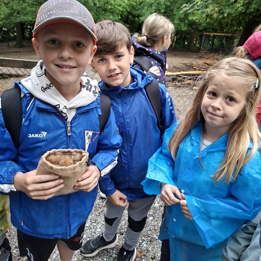 Three children stand in a forest area. A boy in a blue jacket holds a brown bag. Another boy and a girl, both in blue jackets, stand next to him. Behind them, a child and an adult stand near a fence.
