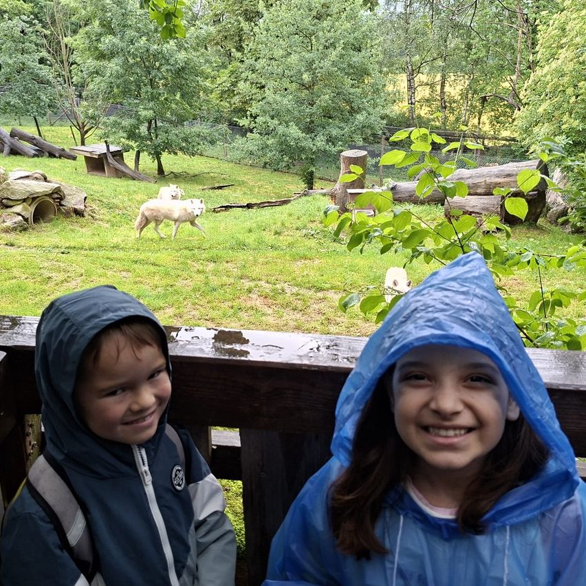 Two children in raincoats are smiling at the camera, with a grassy field and two white wolves in the background.