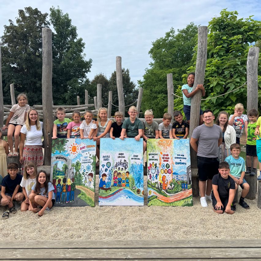 Gruppenfoto von Kindern und Erwachsenen, die in einem Outdoor-Spielplatz stehen und drei bemalte Tafeln präsentieren. Die Kinder und Erwachsenen lächeln und posieren für die Kamera.