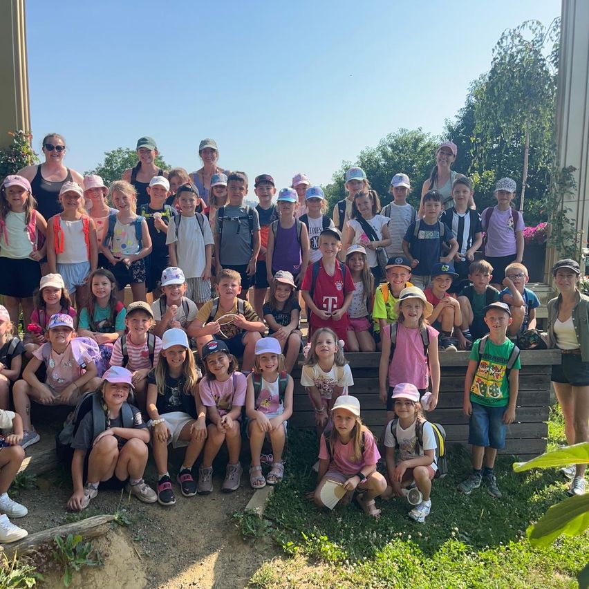 Eine Gruppe junger Kinder, die Mützen und Rucksäcke tragen, versammelt sich auf einem Grasfeld. Sie sitzen und stehen auf einer Holzplattform, möglicherweise für ein Gruppenfoto. Einige von ihnen lächeln und posieren für die Kamera. Dahinter befindet sich eine Betonmauer mit Pflanzen und Bäumen.