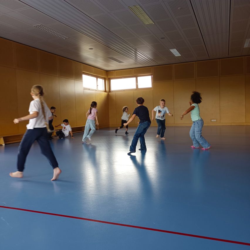 Eine Gruppe von Kindern spielt in einem Innenbereich. Sie sind barfuß und einige laufen, während andere stehen. Der Boden ist blau und hat eine rote Linie an der Seite.