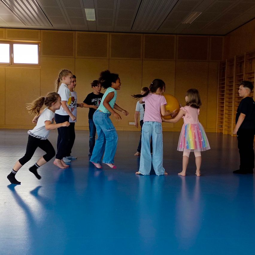 Kinder in einer Turnhalle spielen mit einem Ball, einige halten sich an den Händen, während andere zusehen. Der Raum hat Holzwände und einen blauen Boden.