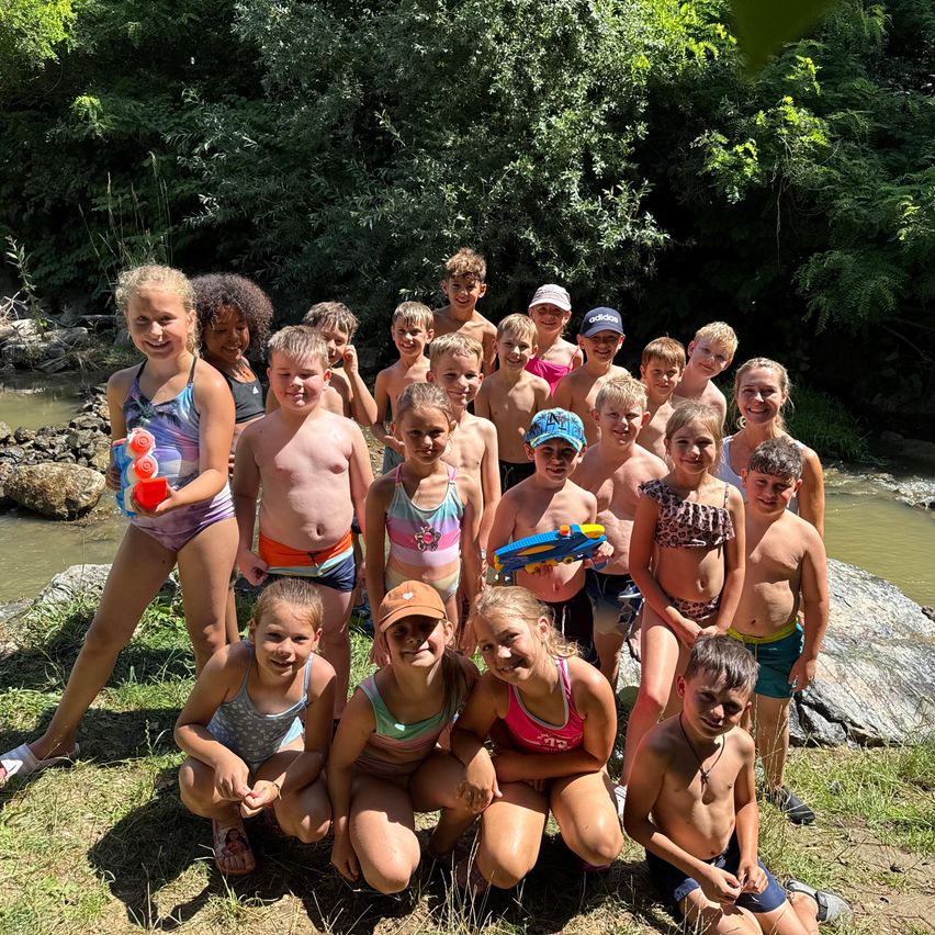 Eine Gruppe von Kindern in Badekleidung steht am Fluss, einige mit Spielzeug, andere in Mützen, in einer natürlichen Umgebung.