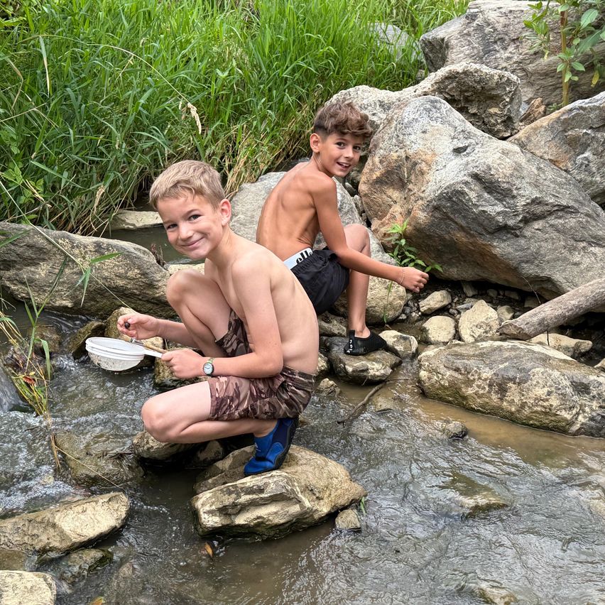 Zwei Jungen sitzen auf Felsen neben einem fließenden Bach, einer hält eine Sieb in der Hand, beide lächeln und genießen die Outdoor-Aktivität.
