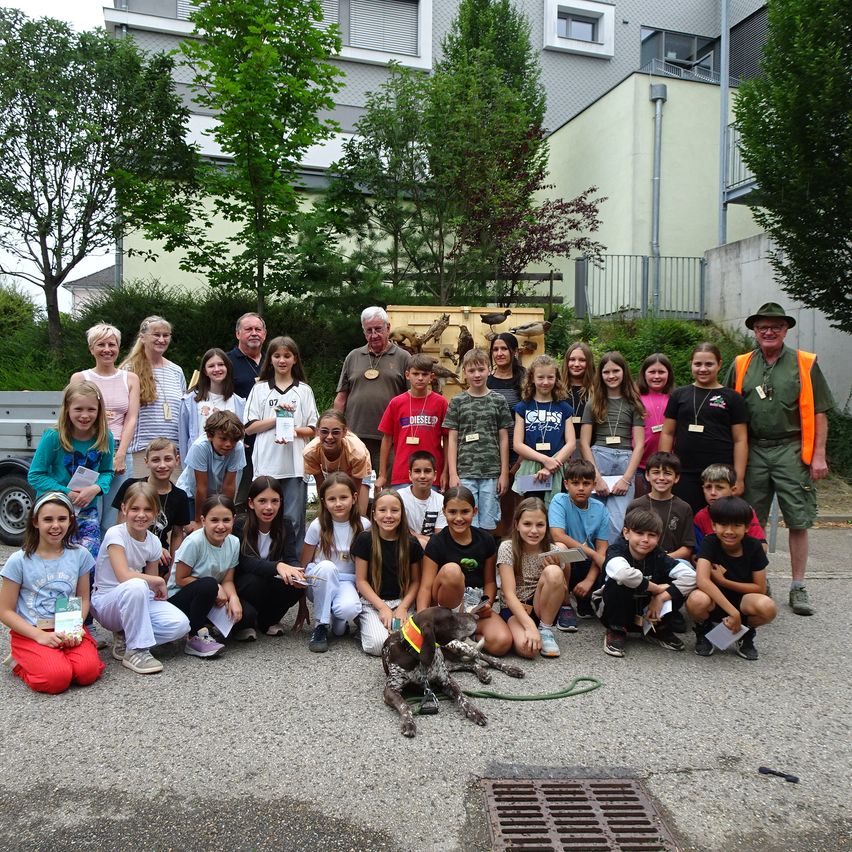 Eine Gruppe von Kindern und Erwachsenen, einschließlich eines Mannes mit einem Hund, posiert für ein Foto vor einem Gebäude mit Bäumen und einer Holzstruktur.