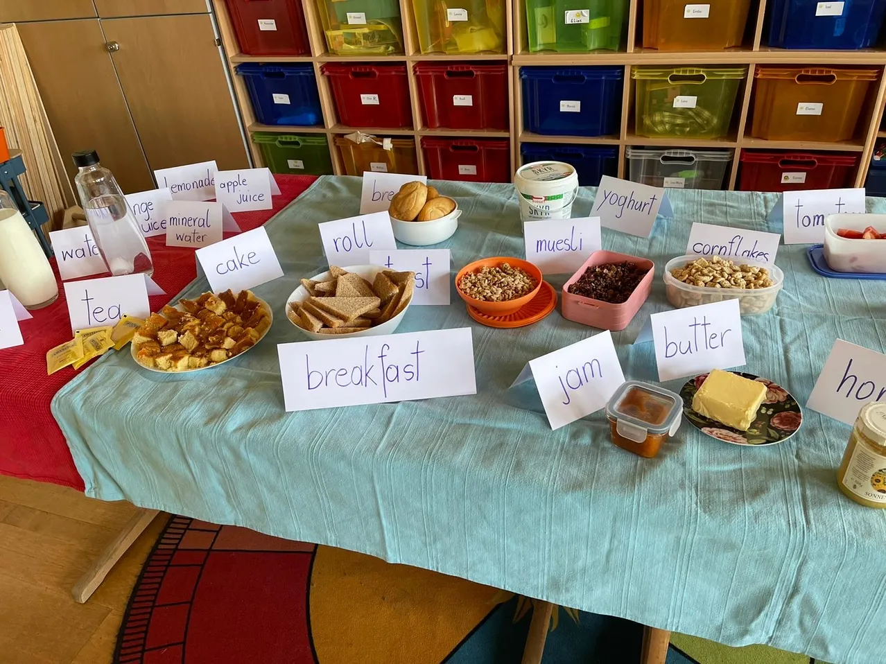 Ein Tisch mit verschiedenen Frühstücksnahrungsmitteln, darunter Brot, Müsli, Obst und Joghurt. Jedes Lebensmittel hat ein Schild mit seinem Namen.