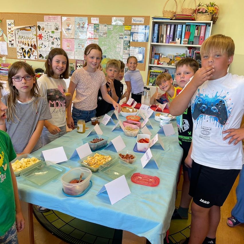 Mehrere Kinder stehen um einen Tisch mit verschiedenen Speisen. Ein Kind isst. Der Tisch ist mit einem blauen Tuch bedeckt und hat mehrere Karten mit Namen.