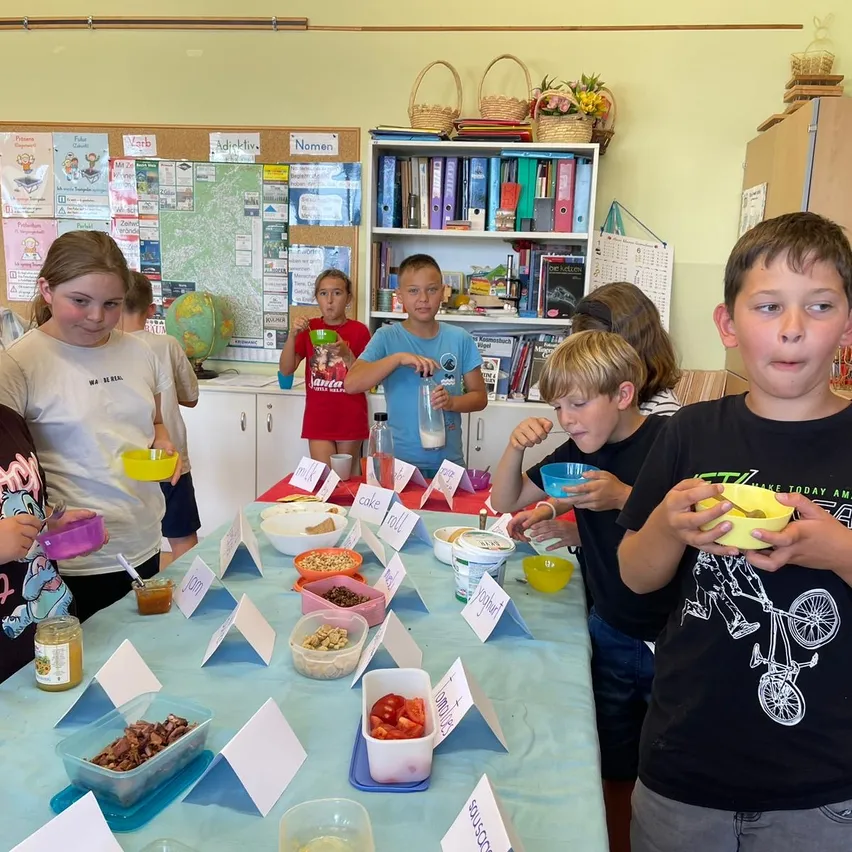 Mehrere Kinder in einem Klassenzimmer versammeln sich um einen Tisch mit verschiedenen Lebensmitteln. Eine Mädchen hält eine Schüssel mit Essen, während andere mit Löffeln essen.