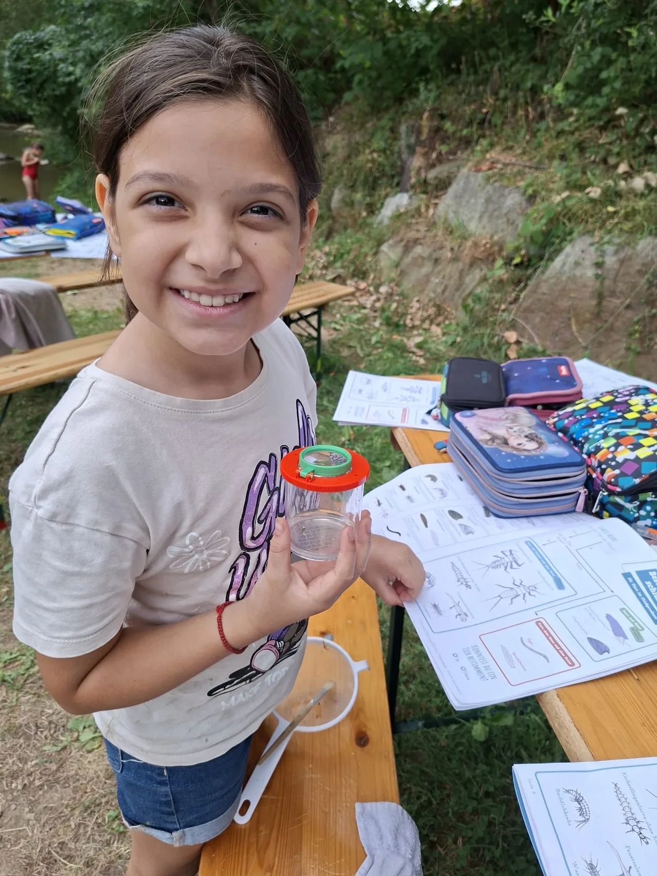 Ein junges Mädchen hält draußen einen Glasbehälter und lächelt. Sie ist in der Nähe eines Picknicktischs mit Papieren, Lunchboxen und einem Fluss im Hintergrund.