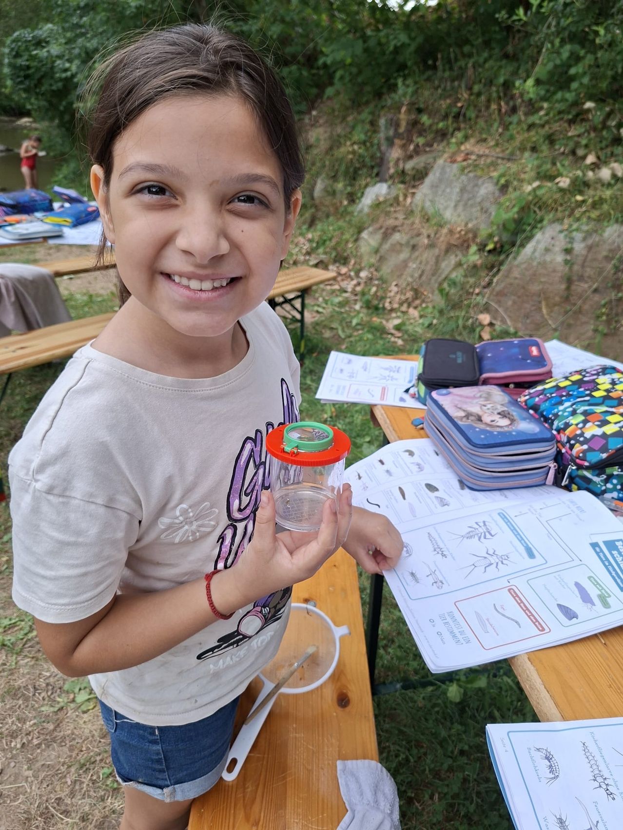 Ein junges Mädchen hält draußen einen Glasbehälter und lächelt. Sie ist in der Nähe eines Picknicktischs mit Papieren, Lunchboxen und einem Fluss im Hintergrund.