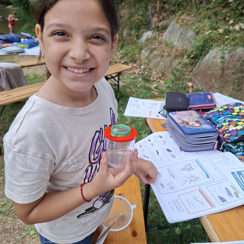 Ein junges Mädchen hält draußen einen Glasbehälter und lächelt. Sie ist in der Nähe eines Picknicktischs mit Papieren, Lunchboxen und einem Fluss im Hintergrund.