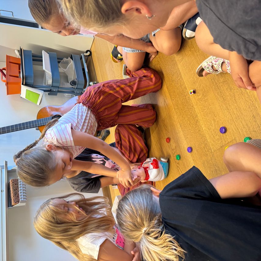 Eine Gruppe junger Mädchen spielt mit Spielzeug auf einem Holzboden, einige sitzend und andere stehend. Bunte Spielzeuge sind verstreut.