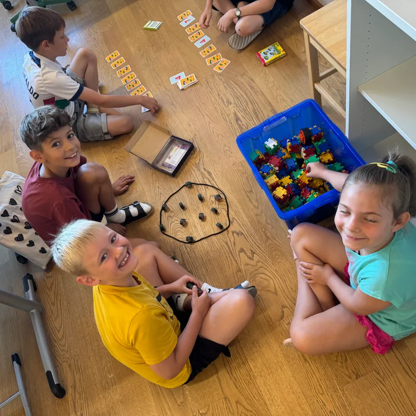 Vier Kinder sitzen auf einem Holzboden und spielen ein Kartenspiel und eine Schachtel mit bunten Bauklötzen. Ein Mädchen lächelt in die Kamera.