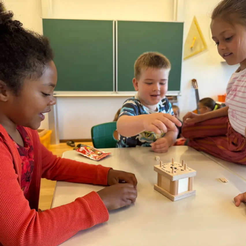 Drei Kinder spielen mit einer Holzbox mit Zapfen auf einem Tisch im Klassenzimmer. Das Mädchen lacht und der Junge ist konzentriert.