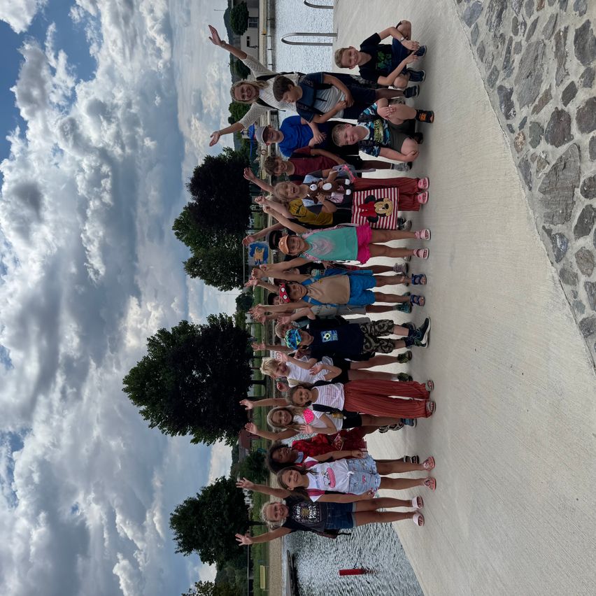 Eine Gruppe von Kindern und Erwachsenen, die zusammenstehen, lächeln und für ein Foto auf einem Pfad am See mit bewölktem Himmel posieren.